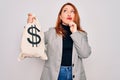 Young beautiful redhead woman holding bag with money and dollar sign over white background serious face thinking about question, Royalty Free Stock Photo