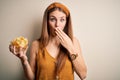 Young beautiful redhead woman holding bowl with potato chips over isolated white background cover mouth with hand shocked with Royalty Free Stock Photo