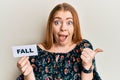 Young beautiful redhead woman holding fall word on paper pointing thumb up to the side smiling happy with open mouth