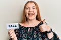 Young beautiful redhead woman holding fall word on paper smiling happy and positive, thumb up doing excellent and approval sign