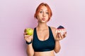 Young beautiful redhead woman holding green apple and cake slice puffing cheeks with funny face Royalty Free Stock Photo