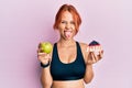 Young beautiful redhead woman holding green apple and cake slice sticking tongue out happy with funny expression Royalty Free Stock Photo