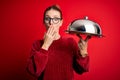 Young beautiful redhead woman holding waitress tray over isolated red background cover mouth with hand shocked with shame for