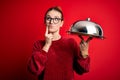 Young beautiful redhead woman holding waitress tray over isolated red background serious face thinking about question, very