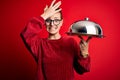 Young beautiful redhead woman holding waitress tray over isolated red background stressed with hand on head, shocked with shame