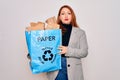Young beautiful redhead woman recycling holding paper bag with cardboard to recycle with a confident expression on smart face Royalty Free Stock Photo