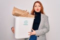 Young beautiful redhead woman recycling holding trash can with cardboard to recycle with a confident expression on smart face Royalty Free Stock Photo
