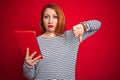 Young beautiful redhead woman using tablet over red isolated background with angry face, negative sign showing dislike with thumbs Royalty Free Stock Photo