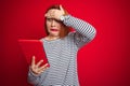 Young beautiful redhead woman using tablet over red isolated background stressed with hand on head, shocked with shame and Royalty Free Stock Photo