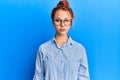 Young beautiful redhead woman wearing casual clothes and glasses over blue background with serious expression on face Royalty Free Stock Photo