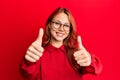 Young beautiful redhead woman wearing casual clothes and glasses over red background approving doing positive gesture with hand, Royalty Free Stock Photo