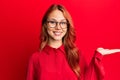 Young beautiful redhead woman wearing casual clothes and glasses over red background smiling cheerful presenting and pointing with Royalty Free Stock Photo