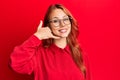 Young beautiful redhead woman wearing casual clothes and glasses over red background smiling doing phone gesture with hand and Royalty Free Stock Photo