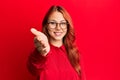 Young beautiful redhead woman wearing casual clothes and glasses over red background smiling friendly offering handshake as Royalty Free Stock Photo