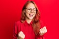 Young beautiful redhead woman wearing casual clothes and glasses over red background very happy and excited doing winner gesture Royalty Free Stock Photo