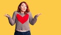 Young beautiful redhead woman wearing casual heart sweater clueless and confused expression with arms and hands raised Royalty Free Stock Photo