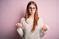 Young beautiful redhead woman wearing casual sweater and glasses over pink background Pointing down looking sad and upset, Royalty Free Stock Photo