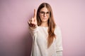 Young beautiful redhead woman wearing casual sweater and glasses over pink background Pointing with finger up and angry Royalty Free Stock Photo