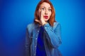 Young beautiful redhead woman wearing denim shirt standing over blue isolated background hand on mouth telling secret rumor, Royalty Free Stock Photo