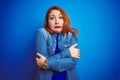 Young beautiful redhead woman wearing denim shirt standing over blue isolated background shaking and freezing for winter cold with Royalty Free Stock Photo