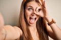 Young beautiful redhead woman wearing diadem make selfie by the camera with happy face smiling doing ok sign with hand on eye Royalty Free Stock Photo
