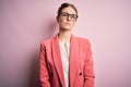 Young beautiful redhead woman wearing jacket and glasses over isolated pink background Relaxed with serious expression on face Royalty Free Stock Photo