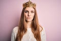 Young beautiful redhead woman wearing queen crown over isolated pink background with serious expression on face Royalty Free Stock Photo