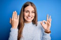 Young beautiful redhead woman wearing wedding ring on finger over blue background doing ok sign with fingers, excellent symbol