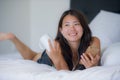 Young beautiful Asian Chinese woman with earpiece listening to music smiling happy lying on bed using internet on mobile phone Royalty Free Stock Photo