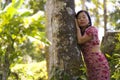 Young beautiful and relaxed Asian Korean woman in elegant summer dress enjoying serene the beauty of nature leaning dreamy on tree