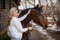 Young beautiful rider woman blonde with long hair in white clothes posing with brown horse Royalty Free Stock Photo