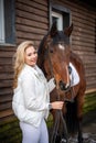 Young beautiful rider woman blonde with long hair in white clothes standing near brown horse on farm Royalty Free Stock Photo