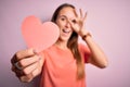 Young beautiful romantic woman holding paper heart shape over isolated pink background with happy face smiling doing ok sign with Royalty Free Stock Photo