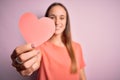 Young beautiful romantic woman holding paper heart shape over isolated pink background with a happy face standing and smiling with Royalty Free Stock Photo