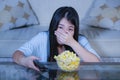 Young beautiful and scared Asian Japanese teenager woman in fear watching horror scary movie at home sofa couch eating popcorn