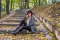 Young beautiful girl model posing in the autumn park among fallen yellow leaves on the old stairs in a hat, coat, jeans and b Royalty Free Stock Photo