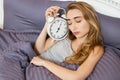 Young beautiful sleepy girl with a closed eyes holds a clock and lies in her bed in the bedroom in the morning Royalty Free Stock Photo