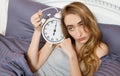 Young beautiful sleepy girl holding a clock and lies in her bed in the bedroom in the morning