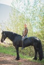 Brunette woman riding dark horse at summer green forest. Royalty Free Stock Photo