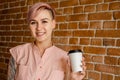 Young beautiful smiling girl holds paper cup of coffee on a brick wall background Royalty Free Stock Photo