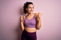 Young beautiful sportswoman with curly hair doing sport drinking glass of water pointing and showing with thumb up to the side