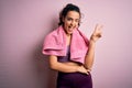 Young beautiful sportswoman with curly hair doing sport using towel over pink background smiling with happy face winking at the