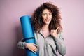 Young beautiful sporty woman with curly hair and piercing holding mat doing yoga happy with big smile doing ok sign, thumb up with