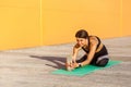 Young beautiful sporty yogi female in black sportwear stretching on the street floor sitting on a mat, looking straight