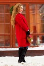 Young, beautiful and stylish blonde hair girl in red coat and with handbag walking through city streets. Womens fashion. Royalty Free Stock Photo