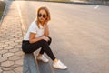 Young beautiful stylish hipster woman with sunglasses in a white polo shirt and black jeans with sneakers sitting on the street Royalty Free Stock Photo