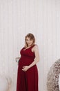 Young beautiful stylish pregnant woman in red long evening dress on white background of child room Royalty Free Stock Photo