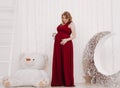 Young beautiful stylish pregnant woman in red long evening dress on white background of child room Royalty Free Stock Photo