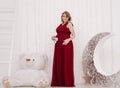 Young beautiful stylish pregnant woman in red long evening dress on white background of child room Royalty Free Stock Photo