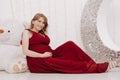 Young beautiful stylish pregnant woman in red long evening dress on white background of child room Royalty Free Stock Photo